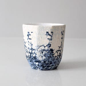 Peut inclure: Une tasse en céramique faite à la main avec un extérieur texturé blanc cassé. La moitié inférieure présente un motif bleu foncé moucheté. La tasse a une forme simple et cylindrique et un intérieur lisse et blanc.