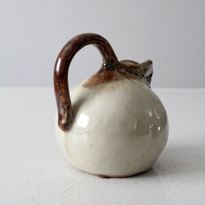 May include: A small, white ceramic pitcher with a brown handle. The pitcher has a rounded body and a slightly flared lip.
