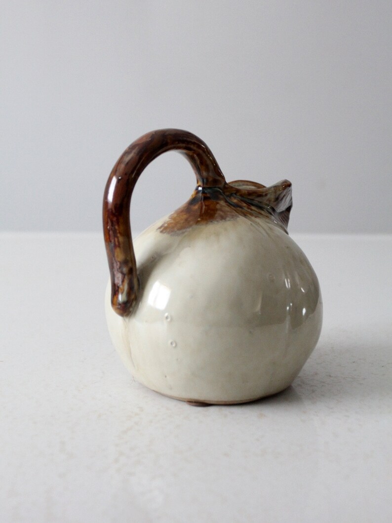 May include: A small, white ceramic pitcher with a brown handle. The pitcher has a rounded body and a slightly flared lip.