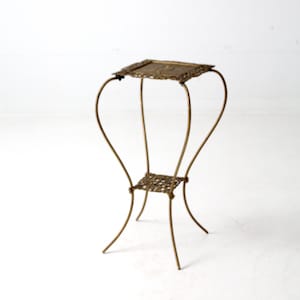Puede incluir: Un soporte para plantas de metal dorado con una parte superior cuadrada y un diseño intrincado. El soporte tiene un estante inferior y patas curvas.