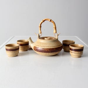 May include: Vintage ceramic tea set with a tan and brown color scheme. The set includes a teapot with a bamboo handle and four matching cups. The teapot and cups have a horizontal brown stripe design.