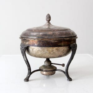 Könnte beinhalten: Silberne Chafing Dish mit runder Basis und drei Beinen. Die Schale hat einen Deckel mit einem dekorativen Knauf.