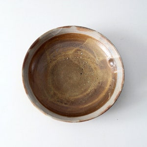 May include: A round ceramic bowl featuring a variegated glaze in shades of earthy brown, tan, and ochre. The interior surface displays subtle tonal variations and small dark speckles, while the rim has a lighter, neutral-toned border.