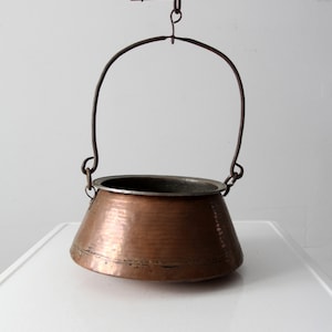 Może przedstawiać: Miedziany garnek z młotkowanym wykończeniem wisi na drewnianym pręcie z metalowym hakiem. Garnek ma szeroką podstawę i wąskie wierzchołek, a jest zawieszony na metalowym pierścieniu.