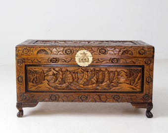 May include: A brown wooden chest with intricate carvings. The chest features a rectangular shape with a detailed relief of sailing ships and floral patterns. A circular emblem adorns the front. The chest rests on four small, curved feet.