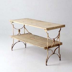 May include: Antique two-tiered table with a light wood top and ornate metal frame. The table has a rectangular shape with a lower shelf. The metal frame is cream-colored and features decorative scrollwork. The table measures approximately 48 inches long.