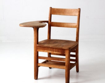 May include: Vintage wooden school chair with attached writing surface. The chair features a slatted back, a rectangular seat, and a small, rounded writing surface on the right side. The wood has a warm, natural finish, and the chair is set against a white background.