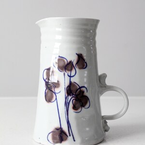 May include: White ceramic pitcher with a blue and brown floral design. The pitcher has a handle and a rounded base.