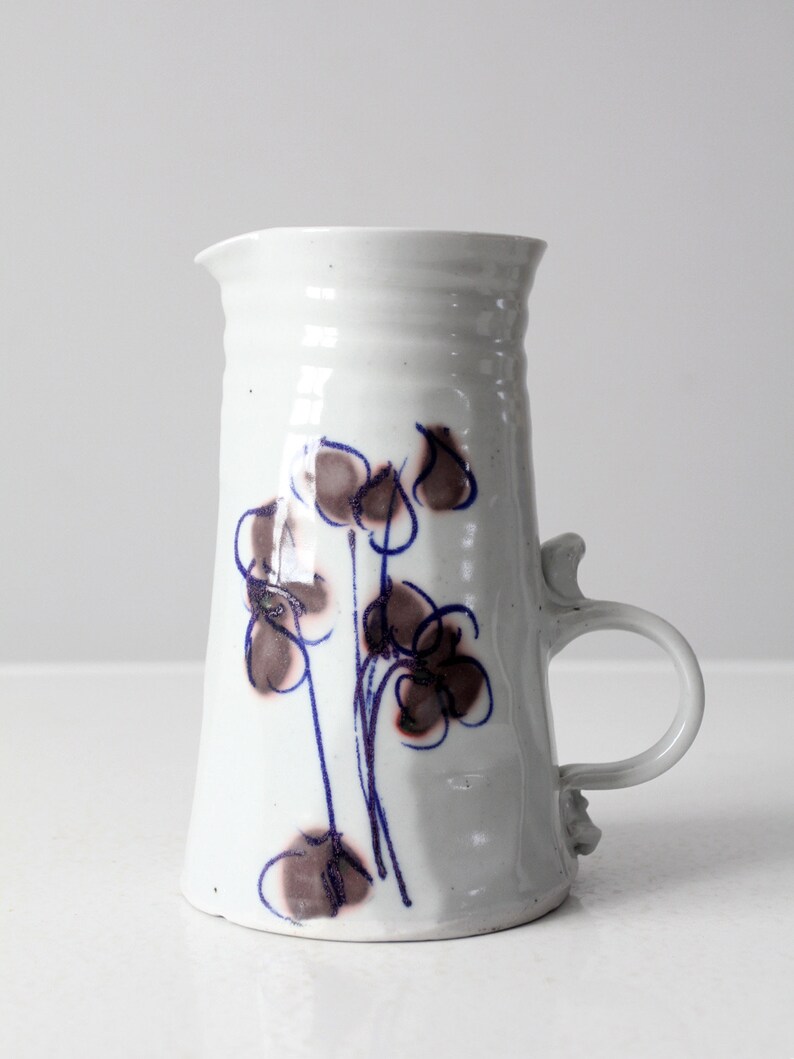 May include: White ceramic pitcher with a blue and brown floral design. The pitcher has a handle and a rounded base.