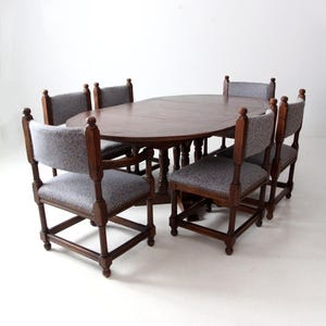 May include: A set of six chairs and a large oval table made of dark wood. The chairs have upholstered seats and backs with a gray patterned fabric. The table has a simple design with a turned pedestal base.
