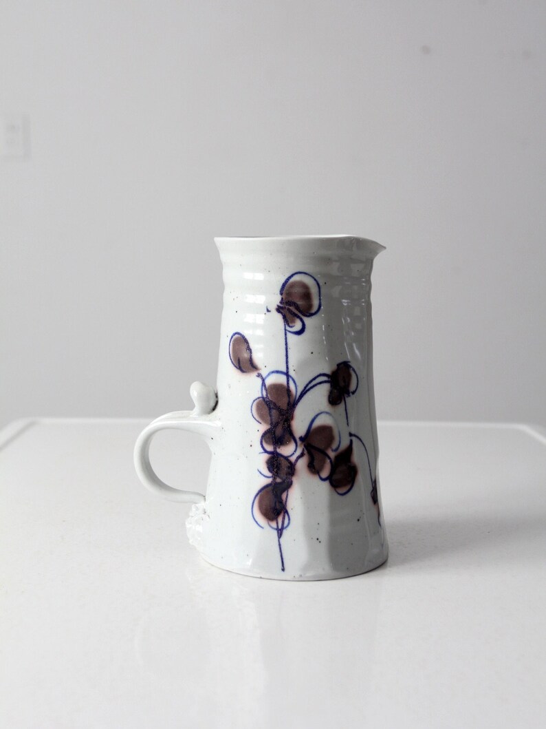 May include: White ceramic pitcher with a brown and blue floral design. The pitcher has a handle and a spout.