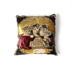 May include: A vintage needlepoint pillow featuring three kittens in a basket. The pillow is made of a gold and black fabric with a red and white floral design. The kittens are depicted in a realistic style with brown and white fur.