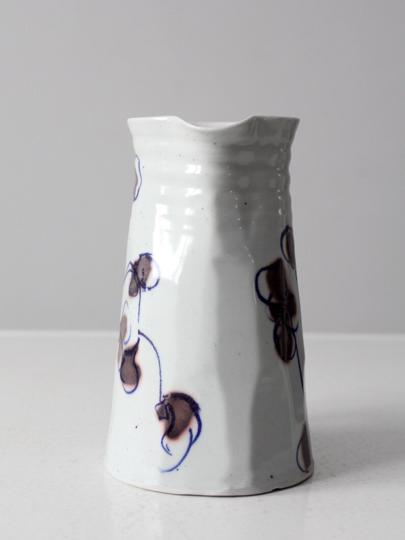 May include: White ceramic pitcher with a blue and brown floral design. The pitcher has a wide mouth and a rounded base.
