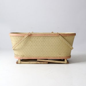 May include: A vintage wicker basket with a light yellow finish and pink trim. The basket has a rectangular shape and is supported by a folding metal frame.