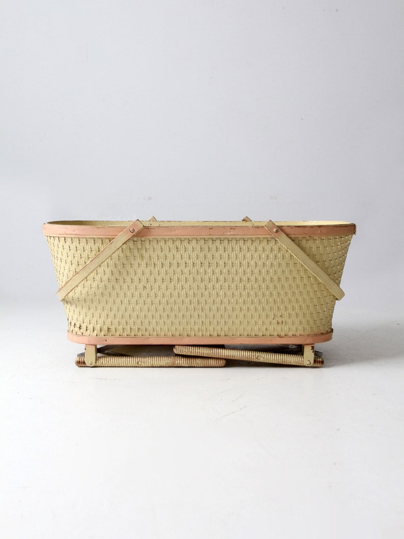 May include: A vintage wicker basket with a light yellow finish and pink trim. The basket has a rectangular shape and is supported by a folding metal frame.