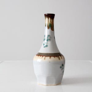 May include: A ceramic vase with a white body and a brown rim. The vase has a slender neck and a wider base with a hexagonal shape. It features green floral accents and a brown and tan band near the top.