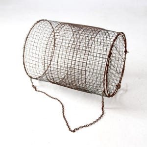 May include: A cylindrical, vintage wire mesh basket with a rusty brown finish. The basket has a chain handle and is likely used for catching or holding items. The design features a series of concentric wire rings.