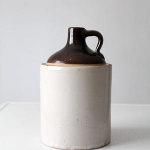 Pode incluir: Uma jarra cilíndrica de grés com um corpo liso em tom branco sujo e uma secção superior contrastante vidrada em castanho escuro. O recipiente inclui uma pequena pega curva fixada ao gargalo, que afunila para uma abertura circular estreita.