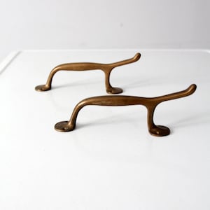 May include: Two antique brass cabinet pulls with a curved design. The pulls have a simple, elegant style and would be a great addition to any home.