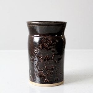 以下が含まれることがあります： 光沢のあるダークブラウンのセラミック製キッチンツールホルダー。ホルダーは、テクスチャ加工されたエンボス加工の花柄のデザインと、明るいベージュ色のベースが特徴です。円筒形は、わずかにフレアした上部を持っています。