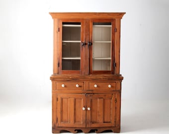 May include: A vintage wooden hutch with glass doors and drawers. The hutch has a light brown finish and features a curved base.