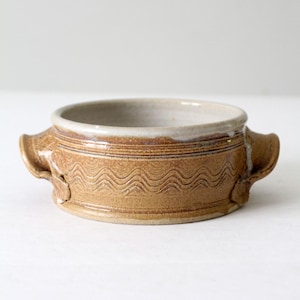 Könnte beinhalten: Eine kleine, braune Keramikschüssel mit zwei Griffen. Die Schüssel hat ein wellenförmiges Muster um den Rand und eine weiße Glasur im Inneren.