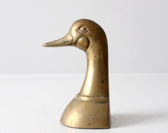 May include: Vintage brass duck head figurine. The duck head is detailed with a long beak, eye, and curved neck. The base is a rounded shape. The brass has a warm, golden tone. A decorative item.