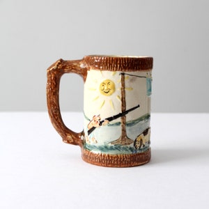 May include: A brown ceramic mug with a tree trunk handle. The mug features a hand-painted scene of a smiling sun, a person holding a rifle, and a dog.