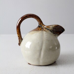 May include: A small, white ceramic pitcher with a brown handle and spout. The pitcher is shaped like a pear and has a glossy finish.