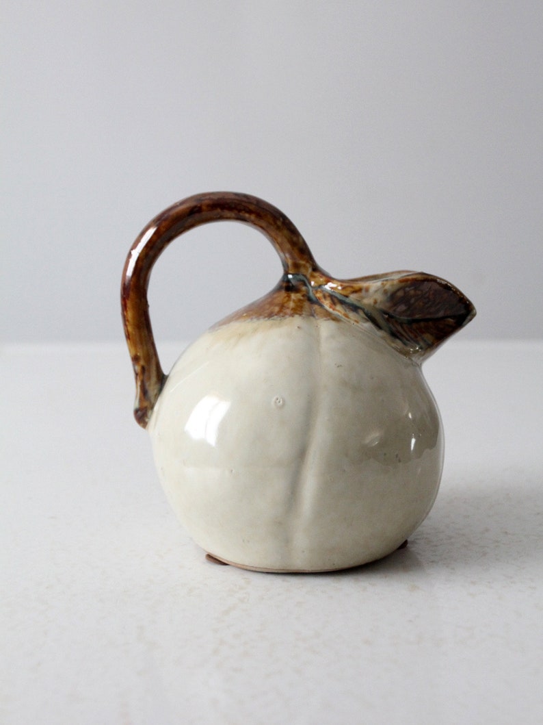 May include: A small, white ceramic pitcher with a brown handle and spout. The pitcher is shaped like a pear and has a glossy finish.