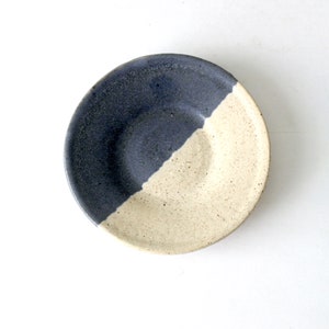 May include: A small, round ceramic dish with a white and blue glaze. The dish is divided in half, with one side being white and the other side being blue.