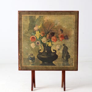 May include: A vintage floral still life painting on a wooden stand. The painting features a dark vase filled with colorful flowers. The stand has a dark brown frame and legs.