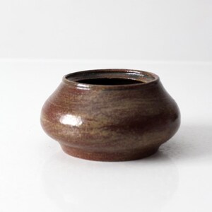 Può includere: Un piccolo vaso in ceramica fatto a mano con un corpo arrotondato e una superficie strutturata marrone scuro. Il vaso ha un'ampia apertura e un bordo leggermente svasato. Il colore è un mix di marrone e beige, con finitura lucida.