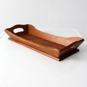 May include: A rectangular wooden serving tray with a raised base and curved sides. The tray has cut-out handles and a rich, warm brown finish. The tray is designed for serving food or displaying items.