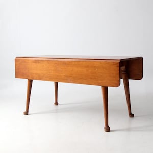 May include: A vintage wooden drop-leaf table with a warm, honey-toned finish. The table features a rectangular top with drop leaves on each side, supported by four slender, tapered legs. The table measures approximately 36 inches long when closed.