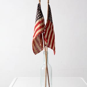 May include: Two small American flags with red and white stripes and a field of stars are displayed in a clear glass bottle. The flags are attached to wooden dowels with decorative tips. The bottle sits on a white surface.