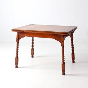 Puede incluir: Una mesa de madera con una hoja abatible y patas torneadas. La mesa está hecha de madera marrón claro y tiene un diseño sencillo.