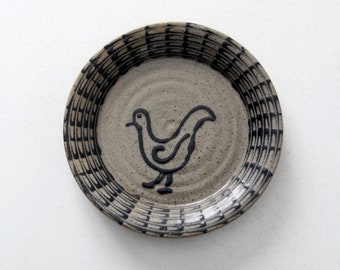 May include: A small, round, ceramic plate with a black and white design. The plate has a black bird design in the center and a black and white geometric pattern around the edge.