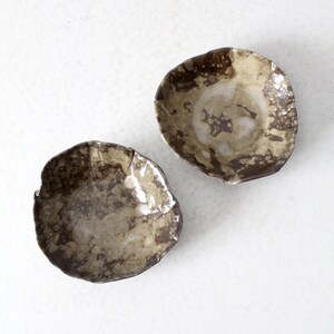 May include: Two small, irregularly shaped ceramic bowls. The bowls have a mottled, organic appearance with shades of brown, tan, and white. The rims are uneven, adding to their rustic charm. They are set against a plain white background.
