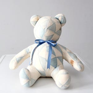 May include: A handmade teddy bear with a patchwork design in cream and light blue. The bear has a blue satin ribbon tied around its neck. The bear is sitting with its arms and legs outstretched.