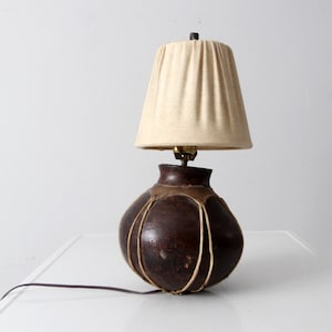 May include: A vintage table lamp with a dark brown, gourd-shaped base and a beige fabric lampshade. The base is adorned with light-colored stitching. The lamp has a brass fitting and a brown electrical cord. The lamp is a decorative lighting fixture.