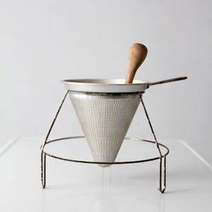 May include: A vintage metal kitchen sieve with a wooden spoon. The sieve has a conical shape with a perforated metal mesh and a handle. It is supported by a wire stand.