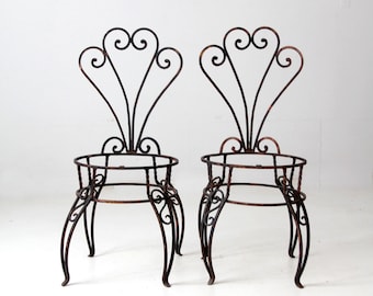 May include: A pair of black wrought iron chairs with ornate scrollwork on the backrests and a circular base. The chairs are in a vintage style and would be perfect for a garden or patio.