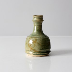 Op de afbeelding: Een kleine, groene keramische vaas met een ronde basis en een smalle hals. De vaas heeft een gespikkelde glazuur met bruine en groene accenten.