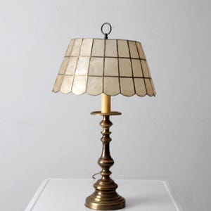 Peut inclure: Lampe de table vintage en laiton avec un abat-jour crème à motif quadrillé. La lampe a un pied orné et un petit anneau en haut. Un élément de décoration intérieure classique.
