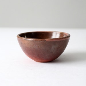 May include: A small, handmade ceramic bowl with a rounded shape. The bowl has a reddish-brown exterior that fades to a lighter, pinkish-red at the base. The interior is a darker, glossy brown. The bowl is likely used for serving small portions of food.