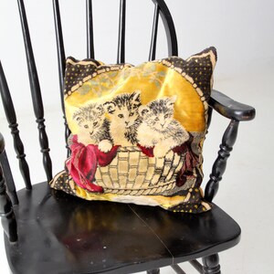 May include: A black wooden chair with a spindle back and a woven tapestry pillow featuring three kittens in a basket. The pillow has a yellow background with brown and red accents.