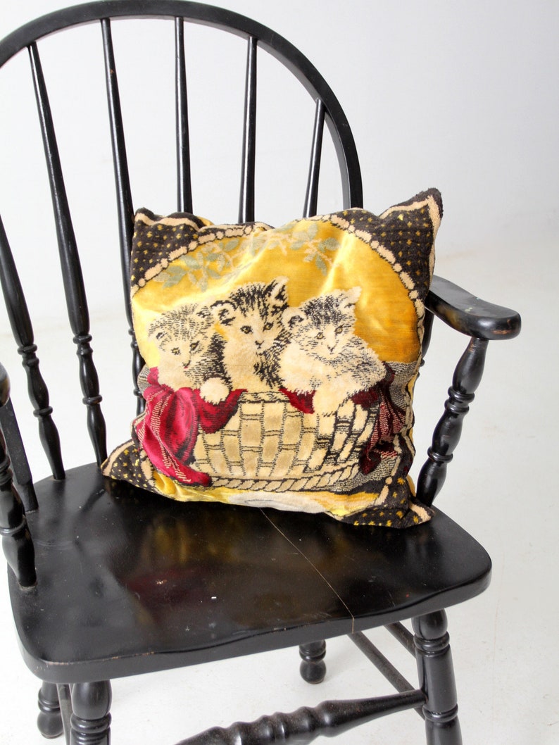 May include: A black wooden chair with a spindle back and a woven tapestry pillow featuring three kittens in a basket. The pillow has a yellow background with brown and red accents.