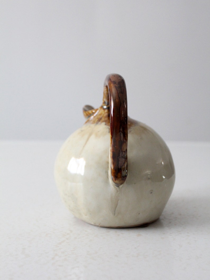 May include: A small, white ceramic pitcher with a brown handle. The pitcher has a rounded body and a slightly flared lip.