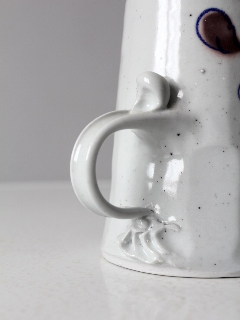 May include: White ceramic pitcher with a speckled glaze and a unique handle. The handle is shaped like a twisted rope and has a small, decorative flower at the base.